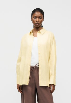 Γυναίκα με πλεξούδες που φοράει ένα ανοιχτό κίτρινο φαρδύ πουκάμισο πάνω από λευκή μπλούζα και καφέ παντελόνι με κορδόνι, μπροστά σε λευκό φόντο.