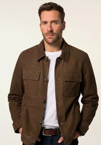 Bruine suède jas met een kraag, knoopsluiting, borstzakken en een gladde textuur, gedragen over een wit t-shirt.