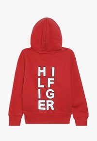 Roter Hoodie mit vorderer Kängurutasche und langen Ärmeln. Auf der Rückseite große weiße Schrift "HILFIGER" in vertikaler Ausrichtung.
