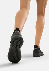 Schwarze Sportschuhe mit strukturierten Sohlen, die ein Slip-On-Design und eine Zuglasche an der Ferse aufweisen, von hinten betrachtet.
