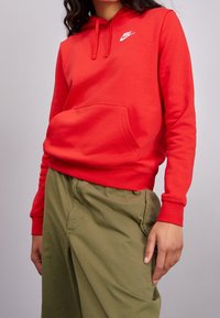 Sweat à capuche rouge en mélange de coton, avec poche frontale et logo Nike blanc. Associé à un pantalon vert clair à coupe décontractée.