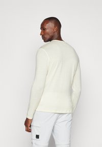 Chemise à manches longues, couleur crème clair, en tissu texturé, avec un col rond et des détails subtils sur la manche gauche. Porte avec un pantalon cargo clair.