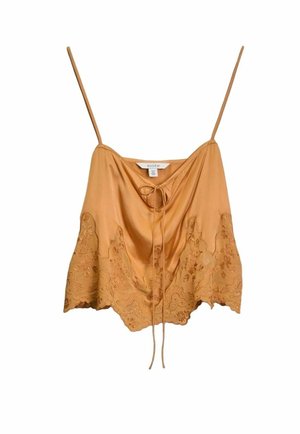 Vrouw met een blauw patroon hoofddoek, oranje kanten crop top, knielange denim shorts en zwarte gespikkelde sandalen, staand tegen een door de zon verlichte muur.