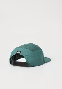 Teal cap with a structured design, mesh panels for ventilation, flat brim, and an adjustable black strap at the back. Brand label attached.