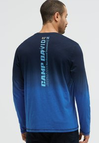 Langarm-T-Shirt in einem Blauverlauf, mit vertikaler weißer Schrift auf dem Rücken und kontrastierenden Nähten am Saum und an den Bündchen.