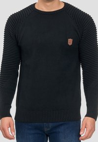 Pull noir en maille avec texture côtelée sur les manches et le corps, col rond classique, et une petite pièce en cuir sur la poitrine.