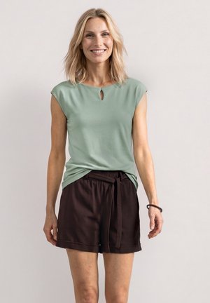 Femme aux cheveux blonds mi-longs portant un haut sans manches vert clair et un short marron foncé, souriant à la caméra.
