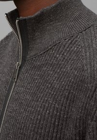 Szary sweter z ribbowanym splotem i wysokim kołnierzem, z metalowym zamkiem błyskawicznym i fakturowanym detalem tkaniny. Zbliżenie uwydatniające kunszt wykonania.