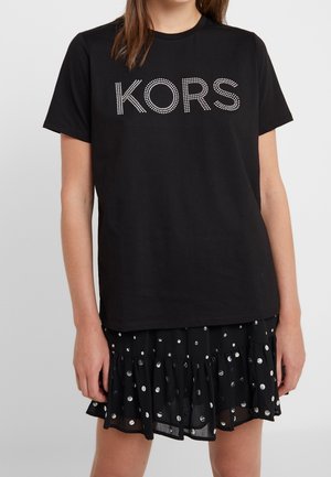 Femme portant un T-shirt noir à manches courtes avec « KORS » en points blancs et une jupe noire avec des motifs circulaires métalliques et un ourlet volanté.