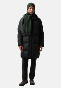 Sort pufferjakke med hætte, grøn strikket hue, gråt halstørklæde og mørke bukser, ledsaget af brune sko med sorte såler.