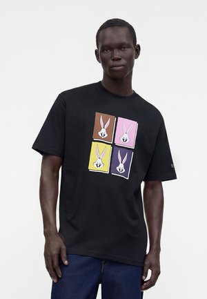 Bărbat purtând un tricou negru cu patru panouri pătrate colorate, fiecare cu chipul lui Bugs Bunny, asortat cu blugi albastru închis.