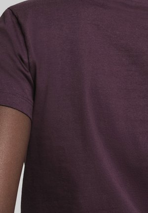 Nærbillede af en mørktonet arm iført en mørkelilla bomuld t-shirt med korte ærmer mod en ensfarvet baggrund.