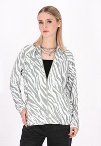 Jachetă cu fermoar cu model de zebră albă și gri, fabricată dintr-un material ușor, cu mâneci lungi și buzunare frontale, purtată peste un top negru.