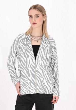 Jachetă cu fermoar cu model de zebră albă și gri, fabricată dintr-un material ușor, cu mâneci lungi și buzunare frontale, purtată peste un top negru.