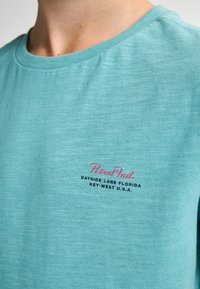 Svetle tyrkysové tričko s krátkým rukávem vyrobené z měkké látky, s růžovým a černým grafickým textovým logem na levé části hrudi.