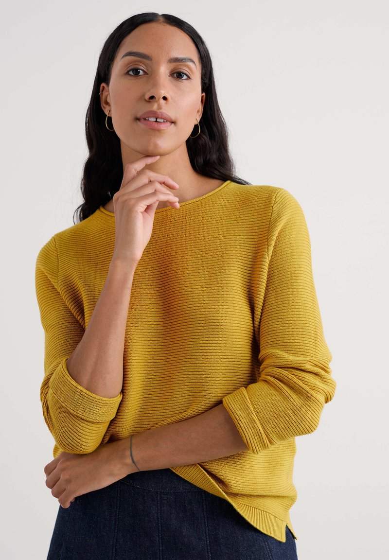 Suéter de punto acanalado amarillo con cuello redondo y puños enrollados, combinado con una falda oscura. Textura clara y sutil detalle de rayas.