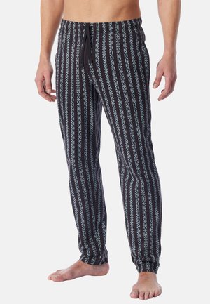 Blootsvoetse man die zwarte en lichtblauwe pyjamabroek met verticale strepen en een koordje in de taille draagt, staand tegen een effen achtergrond.