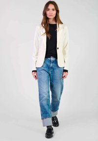 Blazer blanc avec un seul bouton noir, porté par-dessus un top noir. Assorti à un jean large bleu clair avec des ourlets roulés et des chaussures noires.