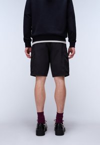 Uomo in piedi di spalle che indossa pantaloncini cargo neri, maglia a maniche lunghe nera, calzini color bordeaux e scarpe eleganti nere su sfondo neutro.