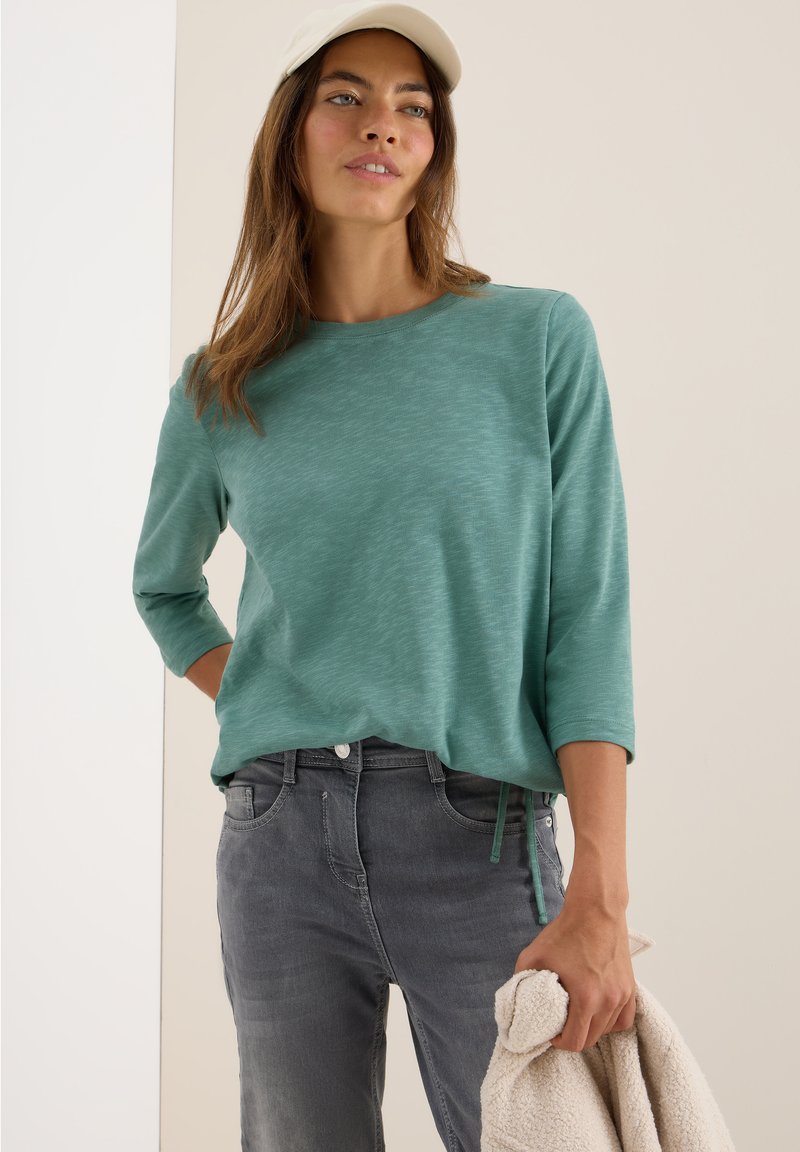 Femme portant un t-shirt à manches longues teal, un jean gris et une casquette beige, tenant une veste en polaire beige dans sa main droite.