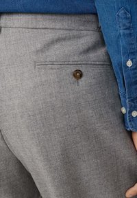 Pantaloni di lana grigi con una tasca posteriore con bottone, indossati con una camicia azzurra a maniche lunghe e bottoni, vista ravvicinata dell'area della vita e dei fianchi.
