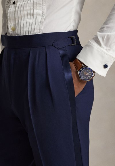 Άτομο που φοράει μπλε σκούρο πλισέ παντελόνι με πλαϊνή αγκράφα και λευκό πουκάμισο, με το χέρι στην τσέπη, με ρολόι χειρός με καφέ δερμάτινο λουράκι ορατό.