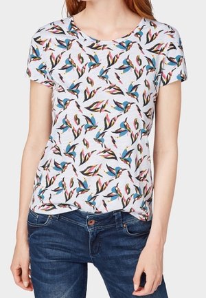Kvinde iført en hvid T-shirt med korte ærmer med farverige fuglemønstre og mørkeblå jeans.