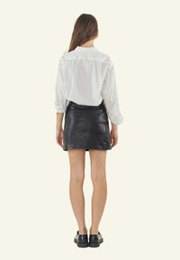 Witte blouse met lange mouwen en opgerolde mouwen, gecombineerd met een zwarte leren minirok. Model draagt zwarte plateau schoenen. Neutrale achtergrond.