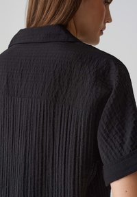 Schwarzes Shirt aus einem strukturierten Stoff mit vertikalen Falten und einem kurzen Kragen. Das gewebte Material hat ein zerknittertes Aussehen.