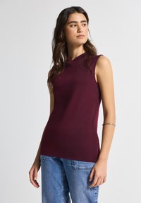 Top sin mangas burdeos con un cuello alto, hecho de una tela suave, estilizado con un corte ajustado, combinado con jeans de color azul claro.