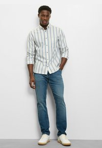 Herrenhemd mit Knopfleiste und vertikalen blau-weißen Streifen, kombiniert mit hellblauen Jeans und weißen Sneakers. Glatter Stoff, lässige Passform.