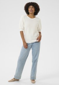 Maglione bianco a maglia con maniche a palloncino corte, abbinato a jeans azzurri a vita alta e mocassini beige intrecciati su uno sfondo neutro.