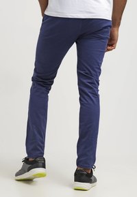 Navy blue stretch chinos with a slim fit. Smooth fabric texture, flat front, and classic back pockets. Worn with black and gray sneakers.
