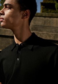 Schwarzes Poloshirt mit kragenförmigem Ausschnitt, drei Knopfleiste und strukturiertem Stoff vor einem hellen Hintergrund.