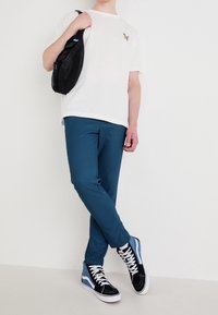 Biała koszulka z krótkim rękawem z małym, kolorowym motylem, turkusowe spodnie i czarne sneakersy za kostkę z niebieskimi akcentami i białymi sznurowadłami.