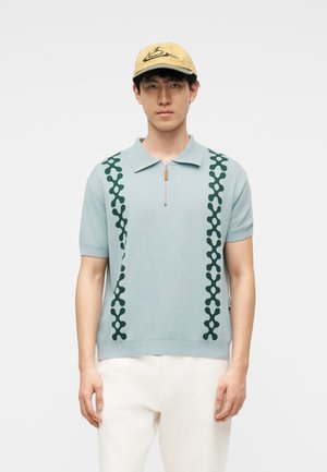 Jeune homme portant un polo bleu clair à manches courtes avec des rayures vertes à motifs, un pantalon blanc et une casquette de baseball beige, debout.