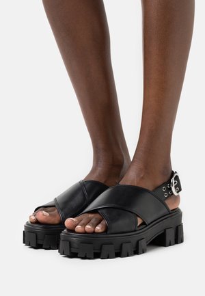 Sandales en cuir noir avec un design à brides croisées, semelle épaisse et chunky, et armature à boucle. Pieds texturés partiellement visibles.