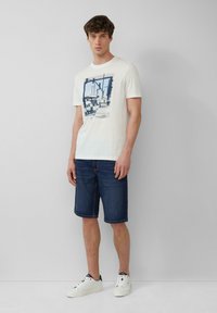 T-shirt en coton blanc avec un imprimé graphique bleu, associé à un short en denim bleu foncé. Mannequin portant des baskets blanches. Fond neutre.