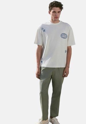 White cotton t-shirt featuring blue graphic accents and logo, paired with light green trousers. Footwear is beige with laces.