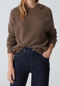 Kobieta w brązowym dzianinowym swetrze z kapturem i ciemnoniebieskich jeansach o wysokim stanie, z jedną ręką w kieszeni, a drugą przy boku.