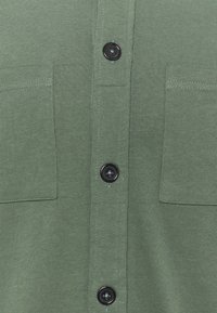 Chemise en coton vert avec devant à boutons, deux poches poitrine et boutons noirs. Le tissu est lisse avec une légère texture.
