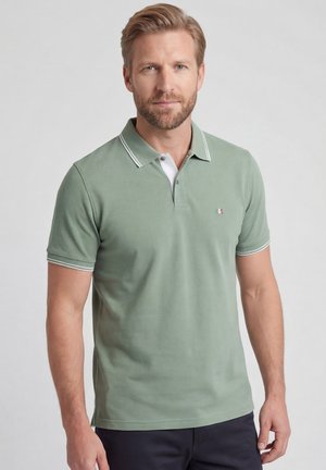 Man met lichtbruin haar en baard, die een lichtgroen poloshirt en donkere broek draagt tegen een effen grijze achtergrond.