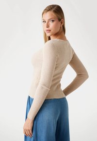 Blonde woman in beige ribbed long-sleeve top and blue high-waist wide-leg jeans poses with hand on hip against white background.