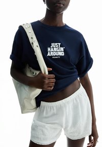 Navy cotton t-shirt with white print stating "JUST HANGIN' AROUND PEANUTS," paired with white embroidered shorts and a cream bag.