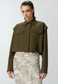 Camicia corta verde oliva con colletto, maniche lunghe e due tasche sul petto, abbinata a una gonna patterned con dettagli metallici.