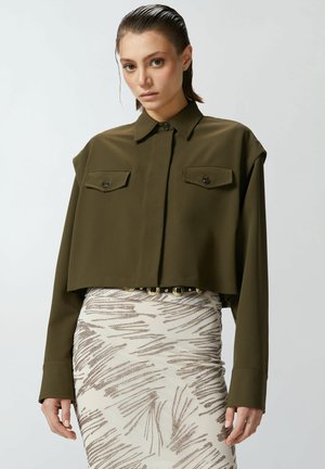 Camicia corta verde oliva con colletto, maniche lunghe e due tasche sul petto, abbinata a una gonna patterned con dettagli metallici.