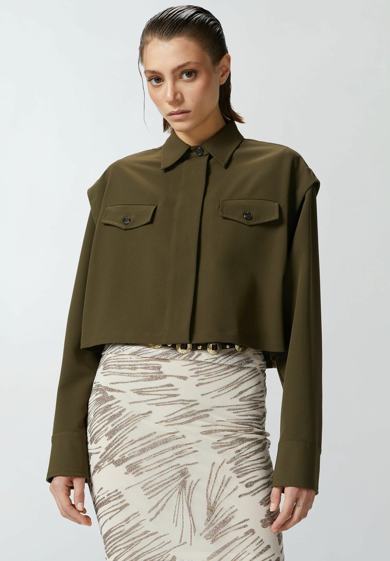 Camicia corta verde oliva con colletto, maniche lunghe e due tasche sul petto, abbinata a una gonna patterned con dettagli metallici.