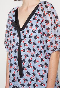 Kobieta ubrana w jasnoniebieską bluzkę z czarno-czerwonym kwiatowym wzorem, czarną lamówką przy dekolcie w kształcie litery V oraz krótkimi bufkami rękawów.