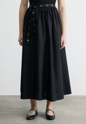 Woman wearing a black high-waisted midi skirt with eyelet belt and black Mary Jane flats, standing on a gray floor against a plain white wall.