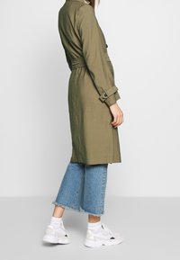 Osoba ubrana w oliwkowy trencz z paskiem, przycięte niebieskie dżinsy z poszarpanymi krawędziami, białe skarpetki i białe trampki, stoi bokiem.